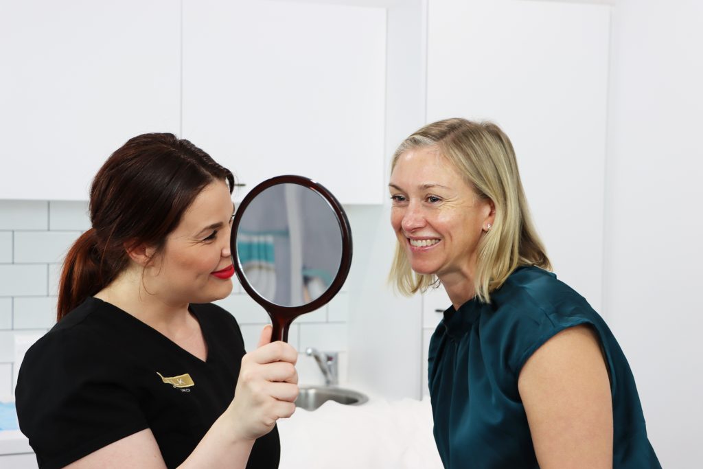 A woman looks at her skin program treatment results in a mirror while another woman beams with a smile beside her